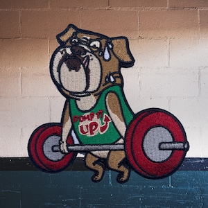 Könnte beinhalten: Ein brauner und weißer Bulldogge, der ein grünes Tanktop mit den Worten "Pump It Up" und einem nach oben zeigenden Pfeil trägt. Der Bulldogge hebt eine Langhantel mit roten und grauen Gewichtsplatten.