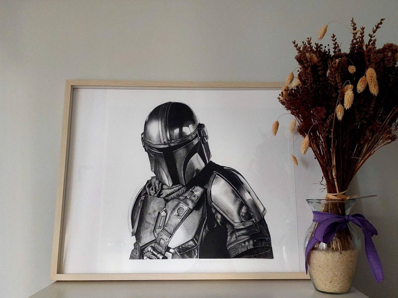 May include: A black and white drawing of a character wearing a helmet and armor, framed in a light wood frame. The drawing is on white paper and is displayed on a white wall.