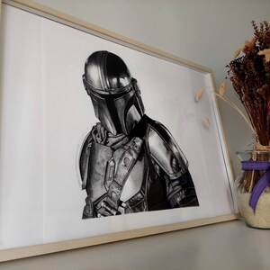 May include: A black and white drawing of a Mandalorian character from the Star Wars franchise. The character is wearing a helmet and armor, and is holding a weapon. The drawing is framed and hanging on a wall.