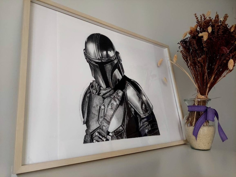 May include: A black and white drawing of a Mandalorian character from the Star Wars franchise. The character is wearing a helmet and armor, and is holding a weapon. The drawing is framed and hanging on a wall.
