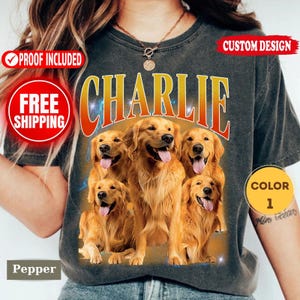May include: A charcoal gray t-shirt featuring a large graphic print of five golden retriever dogs with their tongues out. The word CHARLIE is printed in large, bold, orange-to-yellow gradient letters above the dogs. The shirt has a crew neckline.
