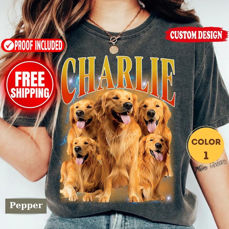 May include: A charcoal gray t-shirt featuring a large graphic print of five golden retriever dogs with their tongues out. The word CHARLIE is printed in large, bold, orange-to-yellow gradient letters above the dogs. The shirt has a crew neckline.