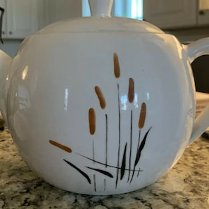 Può includere: Teiera in ceramica bianca con un design di erba alta marrone e nera con steli marroni. La teiera ha un coperchio e un manico.