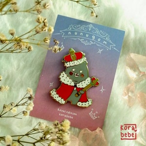 May include: A gold-colored pin featuring a cartoon bear dressed as a king, wearing a red robe and crown, holding a scepter. The pin is on a purple and blue card with the text "nunugom" and "Royal edition korabebe".