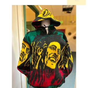 Peut inclure: Un sweat à capuche zippé noir avec un design vibrant avec des accents jaunes, rouges et verts. Le sweat à capuche présente un portrait sur le devant et la capuche, avec le texte "ONE LOVE Collection" visible en haut à gauche.