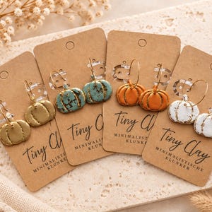 Può includere: Quattro paia di orecchini a forma di zucca su cartoncini marroni con l'etichetta Tiny Clay. Gli orecchini presentano zucche dorate, verde acqua, arancioni e bianche con cerchi in filo metallico dorato. Ogni cartoncino riporta il testo Tiny Clay.
