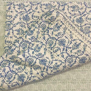 Pode incluir: Uma colcha de algod&atilde;o branco com um padr&atilde;o floral azul. A colcha est&aacute; dobrada ao meio e tem um fundo verde claro.