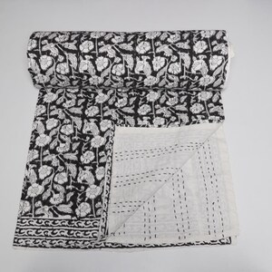 May include: A black and white quilted throw blanket featuring a repeating floral pattern. The top side displays a dense botanical print, while the underside is a solid white fabric with visible parallel running stitch lines throughout the textile.