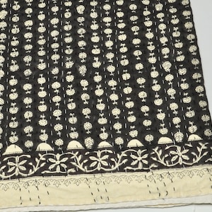 May include: A black textile featuring a repeating pattern of small white floral motifs arranged in vertical columns. The fabric includes a horizontal border with a larger floral vine design and a solid cream-colored edge with white running stitches.