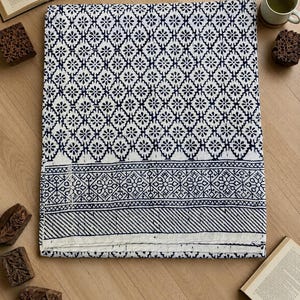 May include: A folded white and navy blue patterned textile, possibly a quilt or blanket, with a repeating floral design. The border features geometric patterns and stripes. Wooden printing blocks and a cup of tea are arranged around the textile on a wooden surface.