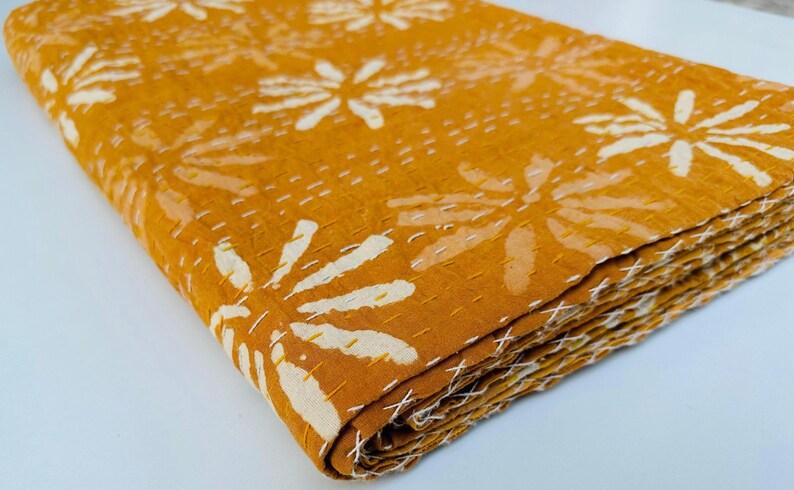 May include: A mustard yellow kantha quilt with a white floral pattern. The quilt is folded in thirds and is made of cotton.