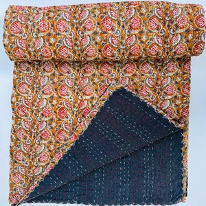 Op de afbeelding: Een omkeerbare quilt met een bloemenpatroon in tinten oranje, rood en wit op een mosterdgele achtergrond. De achterkant is donkergrijs met witte en rode stiksels. De quilt is opgevouwen en opgerold.