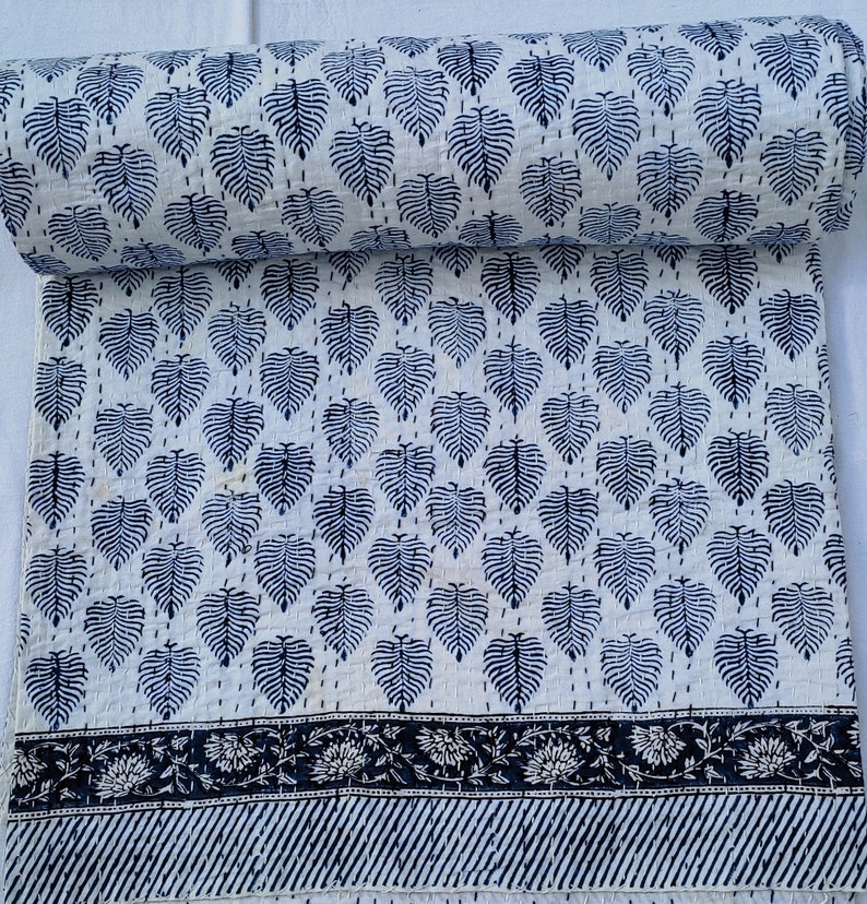 Puede incluir: Colcha kantha blanca con un patr&oacute;n de hojas azules y un borde blanco y negro. La colcha est&aacute; doblada por la mitad y el patr&oacute;n es visible en ambos lados.