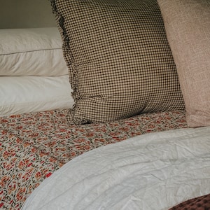 May include: A bed with a white duvet, a patterned throw blanket, and two pillows. One pillow is brown and white gingham with ruffled edges, and the other is a solid brown.