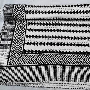 May include: A white cotton quilt featuring a black block-printed pattern. The design includes rows of black triangles, a chevron-style border, and a striped edge. The fabric is finished with visible running stitch quilting details throughout.