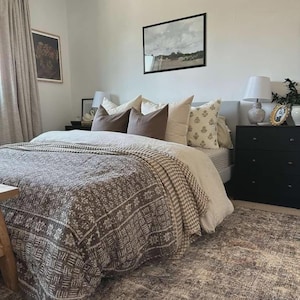 Puede incluir: Un dormitorio con una cama que tiene una colcha marr&oacute;n estampada, un edred&oacute;n de color claro y varias almohadas en tonos marr&oacute;n, crema y blanco. Dos mesitas de noche negras flanquean la cama, cada una con una l&aacute;mpara encima.
