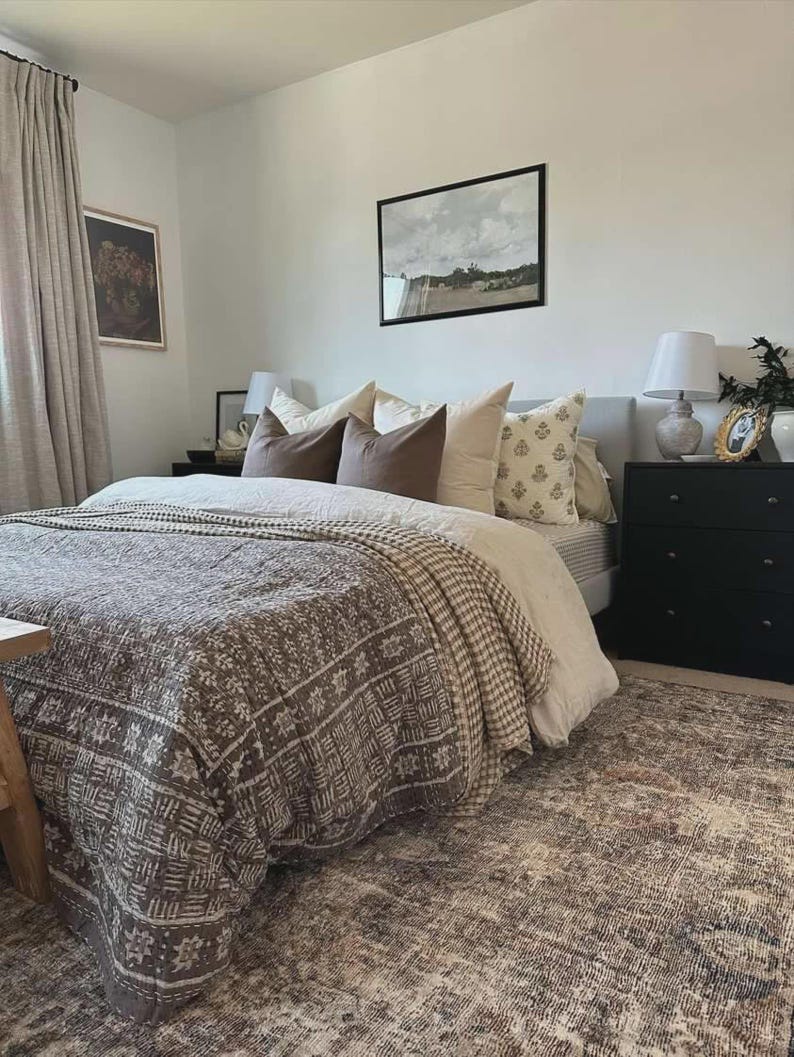 Puede incluir: Un dormitorio con una cama que tiene una colcha marr&oacute;n estampada, un edred&oacute;n de color claro y varias almohadas en tonos marr&oacute;n, crema y blanco. Dos mesitas de noche negras flanquean la cama, cada una con una l&aacute;mpara encima.