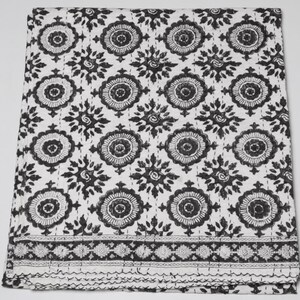 May include: A white quilt featuring a repeating black floral pattern with circular motifs and star-like shapes. The fabric displays visible straight-line stitching throughout, with a distinct horizontal border pattern at the bottom edge.