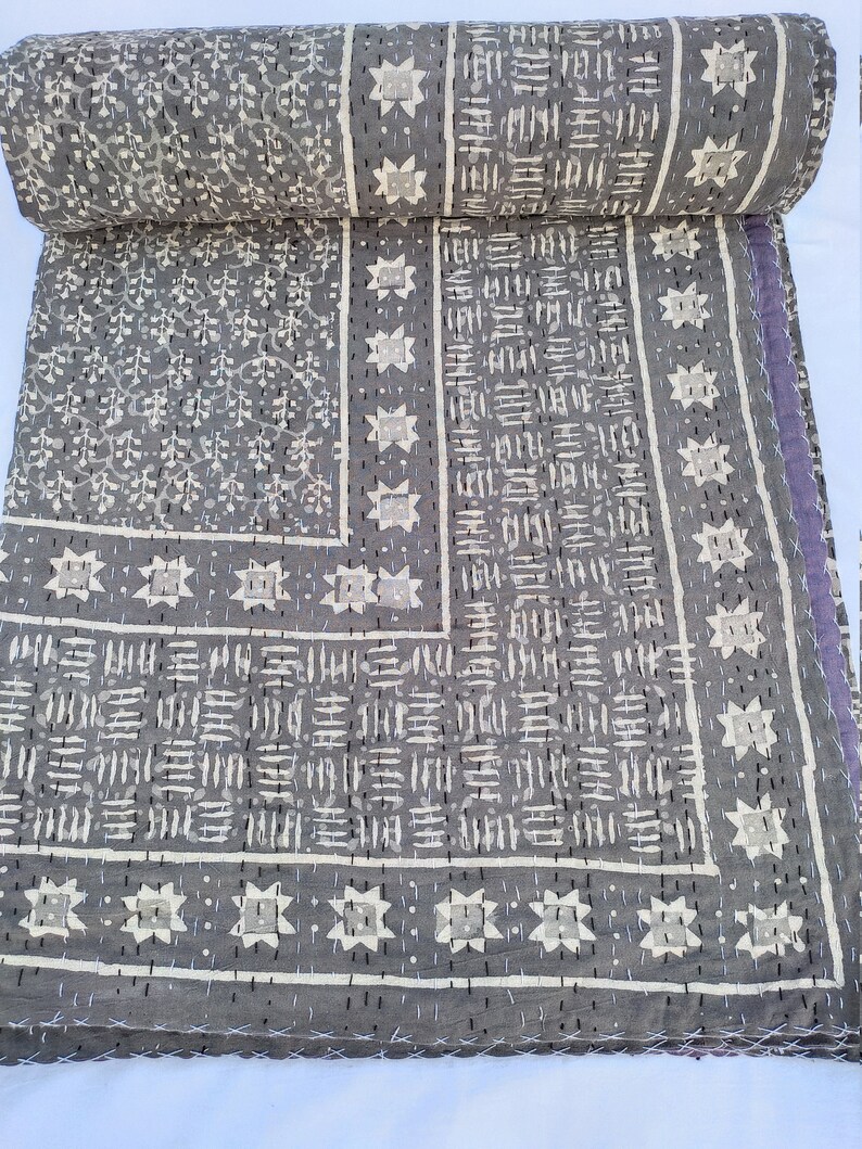 Puede incluir: Una colcha de algod&oacute;n gris con un patr&oacute;n estampado en bloque que incluye motivos florales blancos, formas de estrellas geom&eacute;tricas y l&iacute;neas discontinuas. El tejido presenta bordados visibles de punto hilv&aacute;n en blanco y negro.