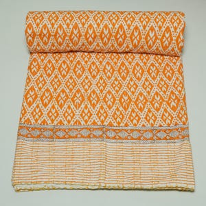 Könnte beinhalten: Eine gefaltete und gerollte orange-weiß gemusterte Steppdecke. Die Steppdecke zeigt ein sich wiederholendes Rautenmuster mit floralen Akzenten. Ein Rand mit einem geometrischen Muster trennt das Hauptdesign von einem gestreiften Abschnitt. Die Steppdecke besteht aus einem weichen Material.