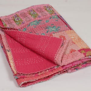 Op de afbeelding: Een gevouwen patchwork deken met diverse stoffen panelen in tinten roze, magenta en perzik. De deken is versierd met ingewikkelde bloemmotieven en gedetailleerd borduurwerk, afgewerkt met zichtbare witte rijgsteek stiksels over het geheel.