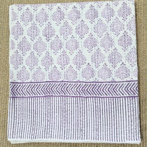 Puede incluir: Una colcha de algodón blanco con un patrón repetitivo de gotas de color púrpura en la sección superior, seguida de una banda horizontal de rayas en zigzag púrpuras y un borde inferior de rayas verticales púrpuras con costuras visibles.