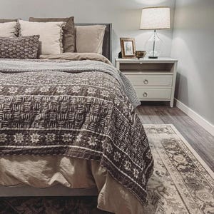 Puede incluir: Una cama con una colcha marr&oacute;n oscuro con un patr&oacute;n geom&eacute;trico blanco, sobre ropa de cama beige lisa. La cama incluye varias almohadas en tonos marr&oacute;n y beige. Una mesita de noche blanca de dos cajones sostiene una l&aacute;mpara y un cuadro.