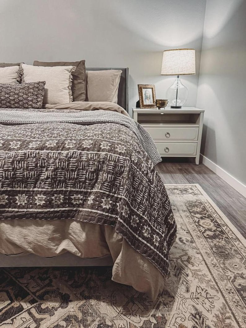 Puede incluir: Una cama con una colcha marr&oacute;n oscuro con un patr&oacute;n geom&eacute;trico blanco, sobre ropa de cama beige lisa. La cama incluye varias almohadas en tonos marr&oacute;n y beige. Una mesita de noche blanca de dos cajones sostiene una l&aacute;mpara y un cuadro.