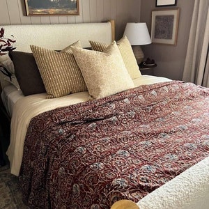 Op de afbeelding: Een bed met een witte hoofdeinde en een rood-witte geruite deken. Het bed heeft verschillende kussens, waaronder twee bruine kussens, twee beige kussens en een gestreept kussen. Het bed staat in een slaapkamer met een houten vloer en een wit tapijt.