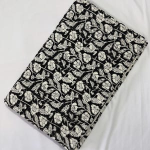 May include: A folded piece of fabric featuring a repeating black and white floral pattern. The design consists of white flowers with detailed petals and leaves arranged on a solid black background, creating a high-contrast botanical print.