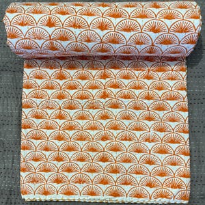 Op de afbeelding: Een opgerold wit en oranje textiel met patroon. Het patroon heeft een herhalend waaierachtig ontwerp in oranje tegen een witte achtergrond. Het textiel is waarschijnlijk een quilt of deken, met zichtbare stiksels langs de randen.