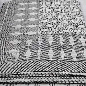 May include: A folded white and black quilt with geometric patterns. The quilt features various designs, including diamond, oval, and striped patterns, all in black stitching on a white background. The quilt is folded, showing multiple layers.