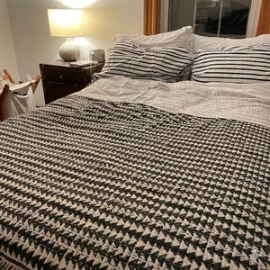 May include: A bed featuring a quilt with a black and white triangular geometric pattern. Two pillows with black and white horizontal stripes are placed at the head of the bed. A white lamp with a textured base sits on a dark wooden nightstand.
