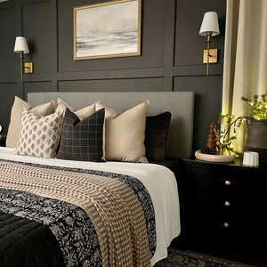 Peut inclure: Une chambre à coucher avec une couette à motifs noir et blanc, une tête de lit grise et une table de chevet noire avec une lampe blanche. Les murs sont gris foncé avec des moulures dorées et un tableau encadré au-dessus de la tête de lit.