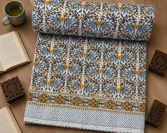 May include: A rolled-up, floral-patterned quilt with a white background, blue and yellow flowers, and a striped border. The quilt is surrounded by books, wooden stamps, and a mug of tea, creating a cozy, inviting scene.