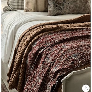 Puede incluir: Una cama blanca con un edredón estampado en marrón y rojo. El edredón tiene una manta de punto marrón texturizada encima. Hay varias almohadas en la cama, incluyendo una almohada estampada con un diseño de cuadros marrones y blancos.