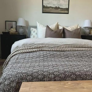 Puede incluir: Una cama con una colcha de patr&oacute;n gris oscuro con costuras blancas, un edred&oacute;n blanco y varias almohadas en tonos crema y marr&oacute;n. Dos mesitas de noche negras con l&aacute;mparas blancas flanquean la cama, con un cuadro de paisaje colgado encima.