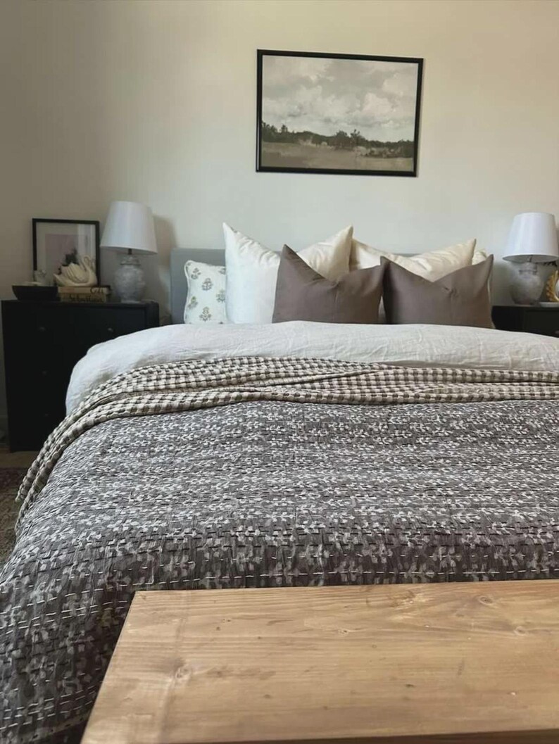 Puede incluir: Una cama con una colcha de patr&oacute;n gris oscuro con costuras blancas, un edred&oacute;n blanco y varias almohadas en tonos crema y marr&oacute;n. Dos mesitas de noche negras con l&aacute;mparas blancas flanquean la cama, con un cuadro de paisaje colgado encima.