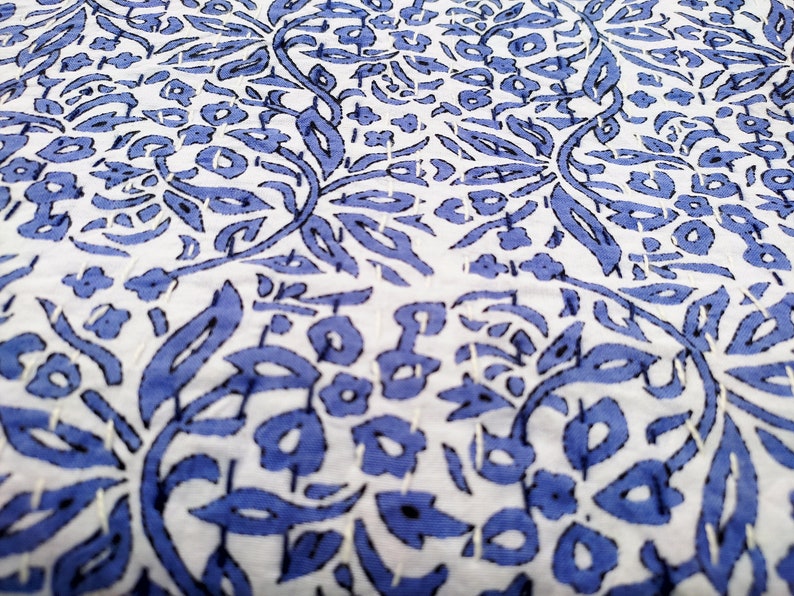 May include: White fabric with a blue floral pattern. The pattern is made up of leaves and flowers. The fabric is quilted with white thread.