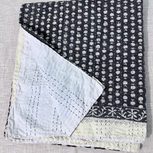 May include: A folded quilt featuring a dark charcoal gray top layer with a repeating white floral pattern and a solid white underside. The fabric displays visible running stitch details throughout, creating a textured appearance with a decorative border.