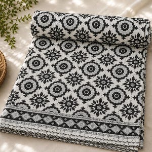 May include: A rolled-up, black and white patterned quilt. The quilt features a repeating floral design and a decorative border. The color scheme is a classic combination of black and white, creating a striking visual contrast. The quilt is folded and rolled, showcasing its intricate design.