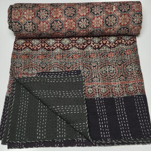 May include: A folded cotton quilt featuring a repeating geometric and floral pattern in shades of red, black, and cream. The reverse side is solid black with white running stitch kantha embroidery arranged in vertical lines across the fabric.