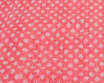 May include: A coral-colored fabric with a repeating pattern of white floral shapes. The design is evenly spaced across the surface, creating a textured appearance. The fabric appears to be a lightweight material, suitable for various textile projects.