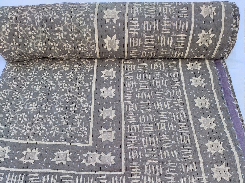 Puede incluir: Una colcha de algod&oacute;n gris con un dise&ntilde;o de patchwork que presenta paneles alternos de motivos florales, patrones de estrellas geom&eacute;tricas y marcas de guiones. La tela est&aacute; detallada con bordados de punto hilv&aacute;n negro visibles.