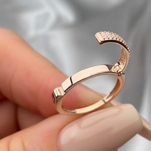Puede incluir: Anillo de oro rosa con una sección superior curvada y con bisagras adornada con pequeñas piedras brillantes. La banda del anillo es lisa y pulida, con un detalle rectangular a cada lado de la bisagra. El anillo es sostenido por una persona.