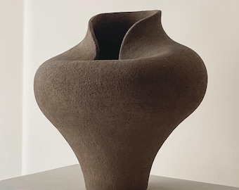 May include: A brown, textured ceramic vase with a unique, organic shape. The vase has a wide, rounded body that tapers to a narrow base and a flared, asymmetrical opening. The neutral color and modern design make it a versatile decorative piece.