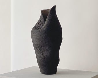 Könnte beinhalten: Eine schwarze, strukturierte Keramikvase mit einer einzigartigen, organischen Form. Die Vase hat eine gefaltete Oberseite und steht auf einem hellgrauen Würfel. Die Oberfläche der Vase hat eine raue, taktile Qualität.