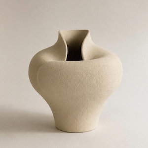 Puede incluir: Un jarrón de cerámica beige con un acabado mate texturizado y una silueta orgánica de curvas distintivas. El recipiente presenta un cuerpo ancho y redondeado que se estrecha hacia una base estrecha y un borde abierto y plegado.