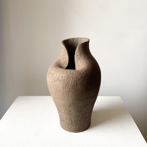 May include: A brown ceramic vase with a rough, textured surface and a distinctively folded, asymmetrical rim. The vessel features a wide, bulbous body that tapers towards a narrow base, showcasing a matte finish and an organic, hand-formed appearance.