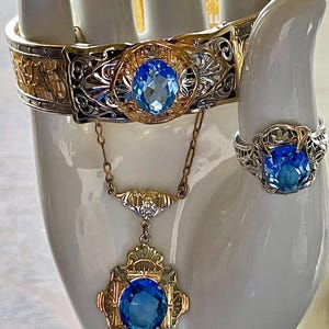 May include: A jewelry set featuring a bracelet, ring, and necklace. The bracelet and ring are silver-toned with gold accents, and the necklace has a gold chain. Each piece has a large, oval, blue gemstone. The jewelry is displayed on a white hand.